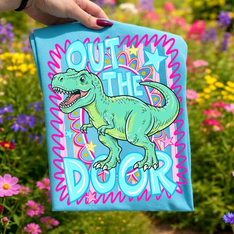 May include: A light blue t-shirt featuring a cartoon Tyrannosaurus Rex in shades of green. The shirt has the words "OUT THE DOOR" in a bubble font, surrounded by pink and blue accents. The shirt is held up by a hand.
