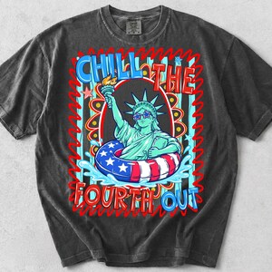 May include: Dark gray t-shirt featuring a graphic of the Statue of Liberty wearing sunglasses and a life preserver with red, white, and blue stripes and stars. The text "CHILL THE FOURTH OUT" is displayed in a colorful font.