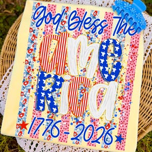 Puede incluir: Una camiseta amarilla clara con las palabras "God Bless The America" en escritura azul. La palabra "America" est&aacute; en rayas rojas y blancas y estrellas azules. Los n&uacute;meros "1776-2026" est&aacute;n en la parte inferior. Una pinza para el pelo azul est&aacute; en la parte superior.