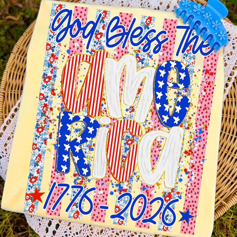 Puede incluir: Una camiseta amarilla clara con las palabras "God Bless The America" en escritura azul. La palabra "America" est&aacute; en rayas rojas y blancas y estrellas azules. Los n&uacute;meros "1776-2026" est&aacute;n en la parte inferior. Una pinza para el pelo azul est&aacute; en la parte superior.