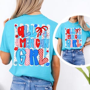 Pode incluir: Uma camiseta azul clara com o texto "ALL AMERICAN GIRL" em vermelho, branco e azul. O texto est&aacute; em uma fonte l&uacute;dica com elementos decorativos como la&ccedil;os, estrelas e fogos de artif&iacute;cio. A camiseta &eacute; de estilo casual.