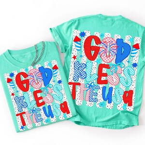 May include: A mint green t-shirt featuring a graphic print with the text "GOD BLESS THE USA" in red, white, and blue patterns. The design includes fireworks, stars, and rocket motifs over a background of vertical black and white dotted stripes.