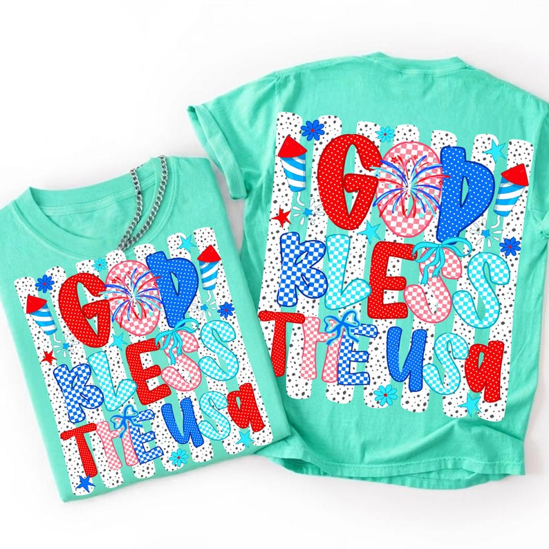 May include: A mint green t-shirt featuring a graphic print with the text "GOD BLESS THE USA" in red, white, and blue patterns. The design includes fireworks, stars, and rocket motifs over a background of vertical black and white dotted stripes.