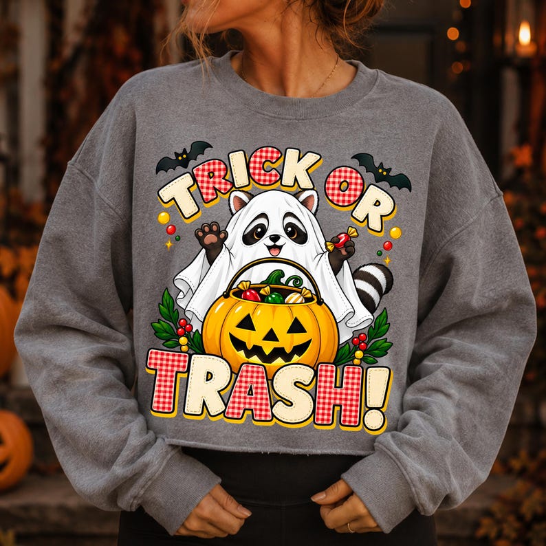 May include: A charcoal grey long-sleeve sweatshirt featuring a graphic of a raccoon dressed as a ghost holding a jack-o'-lantern filled with sweets. The text reads TRICK OR TRASH in red and white checkered lettering, accented by two black bats.