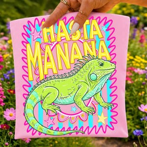 K&ouml;nnte beinhalten: Rosafarbenes T-Shirt mit einer Illustration einer gr&uuml;nen Leguan-Echse und dem gelben Text "HASTA MANANA". Das Design enth&auml;lt Sterne und einen wellenf&ouml;rmigen Rand. Der Leguan hat blaue und gelbe Akzente.
