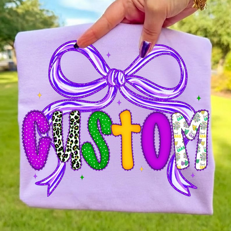 May include: A light purple t-shirt featuring the word "CUSTOM" in a colorful, decorative font. The letters have various patterns and colors, including leopard print, green polka dots, and yellow. A large purple bow with ribbon detail is above the word.