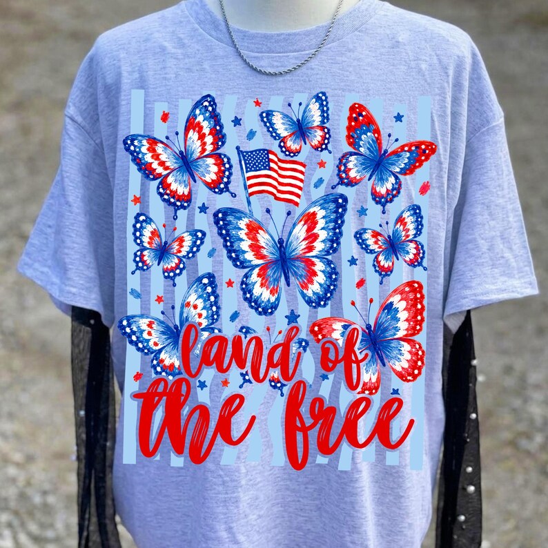 K&ouml;nnte beinhalten: Ein hellgraues T-Shirt mit einem Grafikdruck von mehreren Schmetterlingen mit rot-wei&szlig;-blauen Fl&uuml;gelmustern, einer kleinen amerikanischen Flagge und dem roten Text "land of the free" mittig auf der Vorderseite. Das Design enth&auml;lt kleine blaue Sterne.