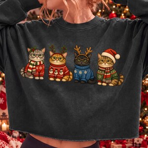 K&ouml;nnte beinhalten: Ein dunkelgraues Sweatshirt mit Rundhalsausschnitt und einem Grafikdruck von vier Katzen, die Brillen und weihnachtliche Kleidung tragen. Die Katzen sind mit Pullovern, Schals und festlichen Kopfbedeckungen wie Rentiergeweihen und einer Weihnachtsm&uuml;tze bekleidet.