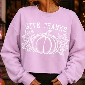 Puede incluir: Una sudadera de manga larga de color morado claro con un estampado gr&aacute;fico blanco de una calabaza rodeada de hojas de oto&ntilde;o. El texto GIVE THANKS est&aacute; impreso en blanco sobre la calabaza. La sudadera tiene cuello redondo y dobladillo sin rematar.