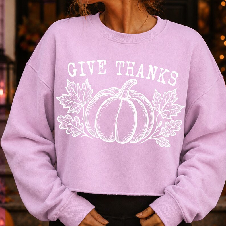 Puede incluir: Una sudadera de manga larga de color morado claro con un estampado gr&aacute;fico blanco de una calabaza rodeada de hojas de oto&ntilde;o. El texto GIVE THANKS est&aacute; impreso en blanco sobre la calabaza. La sudadera tiene cuello redondo y dobladillo sin rematar.