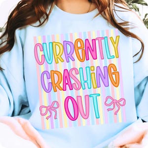 Pode incluir: Camisola azul clara com a frase "CURRENTLY CRASHING OUT" em letras coloridas. O texto est&aacute; sobre um fundo de riscas verticais pastel, com la&ccedil;os cor-de-rosa em ambos os lados da palavra "OUT".
