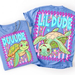 May include: Two light blue t-shirts featuring turtle designs. One shirt displays "DUUUDE" and the other "LIL DUDE" in turquoise bubble letters. The turtles are green and yellow with pink and yellow details.