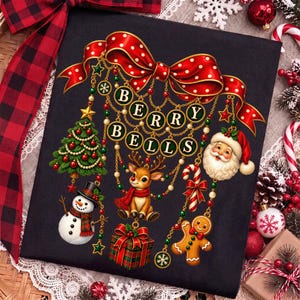 Op de afbeelding: Een zwart t-shirt met een afbeelding van een grote rode strik met stippen en hangende gouden kettingen. Aan de kettingen hangen ornamenten waaronder een kerstboom, een sneeuwpop, een rendier, een cadeau, een zuurstok, een peperkoekmannetje en de Kerstman. De tekst luidt BERRY BELLS.