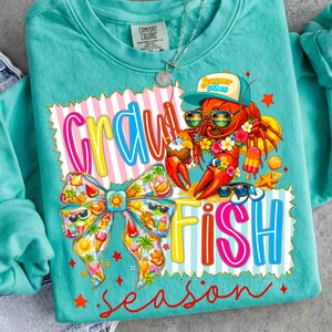 May include: Teal Comfort Colors sweatshirt with a vibrant graphic. The design includes a lobster wearing sunglasses and a hat, with the words "Crawfish Season" in a playful font. A bow and summer elements complete the design.