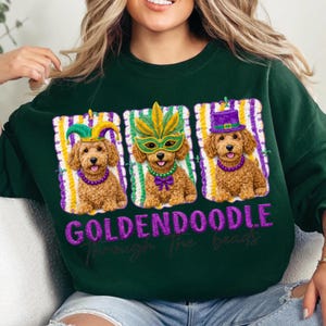 May include: Dark green sweatshirt featuring three Golden Doodle dogs wearing Mardi Gras attire. Each dog is framed with purple, yellow, and white stripes. The text "GOLDENDOODLE Through the beads" is printed in purple.