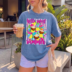 May include: A blue short-sleeve t-shirt featuring a graphic print of coconuts, lemon slices, cherries, and ice cubes over a pink and white striped background. The text "hello sweet summer" is printed in a bright pink script font on the shirt.