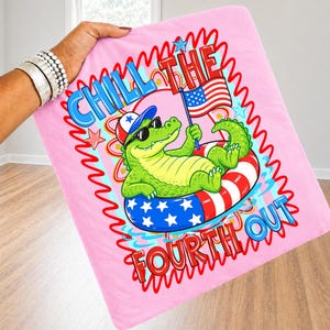 K&ouml;nnte beinhalten: Rosa Decke mit dem Thema 4. Juli. Die Decke zeigt ein Cartoon-Alligator mit Sonnenbrille und patriotischem Hut, der in einem rot-wei&szlig;-blauen Schwimmreifen schwimmt. Der Text auf der Decke lautet "Chill the Fourth Out."