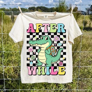 K&ouml;nnte beinhalten: Cremefarbenes T-Shirt mit den Worten "AFTER WHILE" in bunten Buchstaben. Ein Cartoon-Alligator mit Rucksack steht vor einem schwarz-wei&szlig; karierten Hintergrund, umgeben von Blumen.