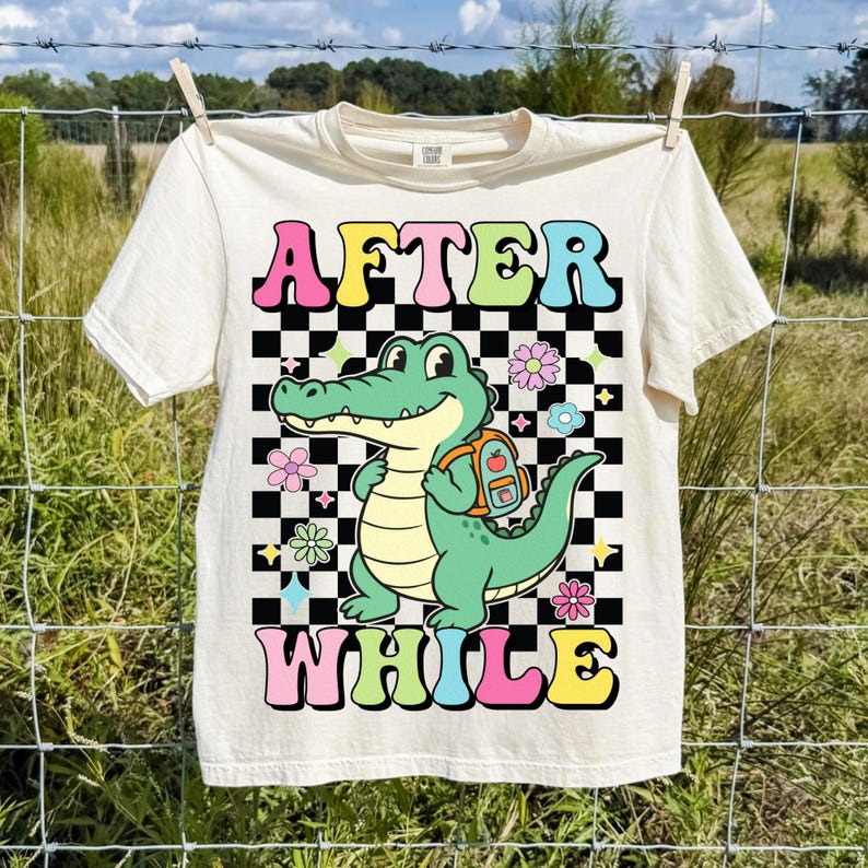 K&ouml;nnte beinhalten: Cremefarbenes T-Shirt mit den Worten "AFTER WHILE" in bunten Buchstaben. Ein Cartoon-Alligator mit Rucksack steht vor einem schwarz-wei&szlig; karierten Hintergrund, umgeben von Blumen.