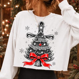 K&ouml;nnte beinhalten: Ein wei&szlig;es, lang&auml;rmeliges, bauchfreies Sweatshirt mit einer grafischen Illustration eines stilisierten Weihnachtsbaums. Der Baum ist mit schwarz-wei&szlig;en Ornamenten, einer roten Kugel, einer gro&szlig;en roten Schleife und einem Stern verziert.
