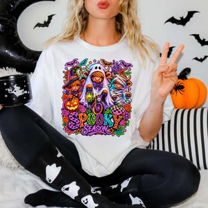 Może przedstawiać: Biały t-shirt z kolorową grafiką przedstawiającą trzy krowy rasy Highland w kostiumach na Halloween: czarownicy, ducha i mumii, z napisem "SPOOKY SEASON" wykonanym żywymi, wzorzystymi literami w otoczeniu nietoperzy i dyń.