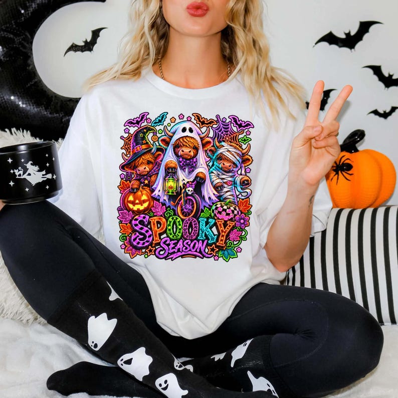 Może przedstawiać: Biały t-shirt z kolorową grafiką przedstawiającą trzy krowy rasy Highland w kostiumach na Halloween: czarownicy, ducha i mumii, z napisem "SPOOKY SEASON" wykonanym żywymi, wzorzystymi literami w otoczeniu nietoperzy i dyń.