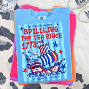 Pu&ograve; includere: Una maglietta azzurra con la scritta "Spilling the Tea Since 1773" e una grafica di una nave con vele rosse, bianche e blu. La maglietta &egrave; piegata sopra altre magliette colorate, tra cui rosa e arancione.