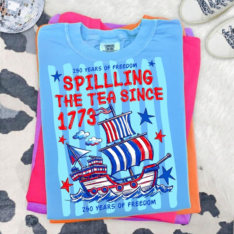 Pu&ograve; includere: Una maglietta azzurra con la scritta "Spilling the Tea Since 1773" e una grafica di una nave con vele rosse, bianche e blu. La maglietta &egrave; piegata sopra altre magliette colorate, tra cui rosa e arancione.