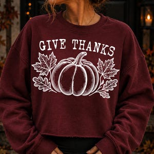 Puede incluir: Una sudadera granate de manga larga con cuello redondo que presenta un estampado gr&aacute;fico blanco de una calabaza centrada entre dos hojas de arce. El texto GIVE THANKS aparece en una fuente serif blanca sobre la ilustraci&oacute;n central.
