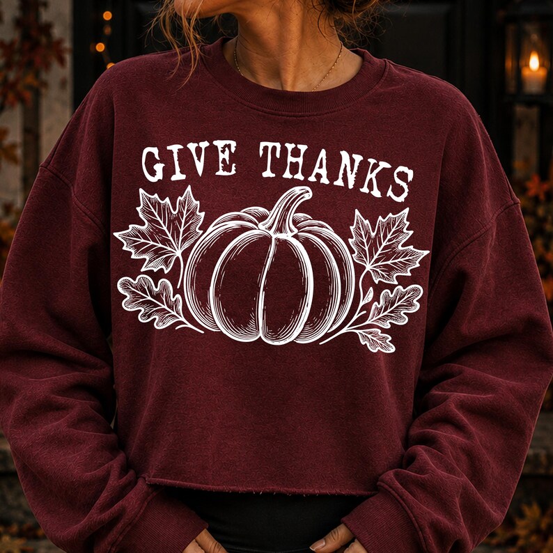 Puede incluir: Una sudadera granate de manga larga con cuello redondo que presenta un estampado gr&aacute;fico blanco de una calabaza centrada entre dos hojas de arce. El texto GIVE THANKS aparece en una fuente serif blanca sobre la ilustraci&oacute;n central.