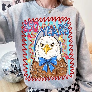 Pu&ograve; includere: Una felpa grigia con una grafica di un'aquila con un papillon blu e la scritta "250 YEARS" in rosso e blu. Il design include stelle e un bordo rosso ondulato, creando un tema patriottico.