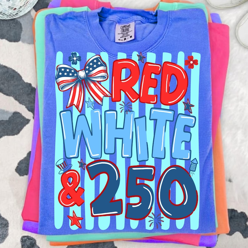 Puede incluir: Una pila de camisetas coloridas, con una camiseta azul en la parte superior con el texto "RED WHITE & 250" en rojo y azul. El dise&ntilde;o incluye un lazo patri&oacute;tico, estrellas y fuegos artificiales. Las camisetas son en tonos de rosa, verde y naranja.