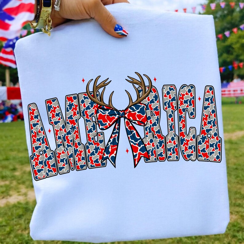 Pu&ograve; includere: Felpa bianca con la scritta "AMERICA" in un design patriottico. Le lettere sono riempite con un motivo mimetico rosso, bianco e blu. Un fiocco e delle corna sono al centro della parola. La felpa &egrave; tenuta da una persona.