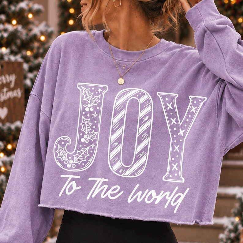 May include: A light purple long-sleeve cropped sweatshirt featuring the white text JOY with holly and candy cane patterns, and the text To The world underneath. The garment has a crew neckline and a raw, unfinished hemline at the waist.