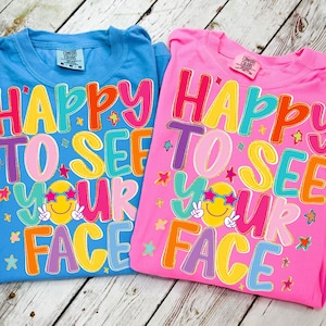 以下が含まれることがあります： 青とピンクの2枚のTシャツ。カラフルなバブルレターで「HAPPY TO SEE YOUR FACE」と書かれています。各シャツには、サングラスとピースサインをした笑顔の顔が描かれています。
