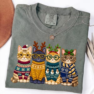 May include: A sage green t-shirt featuring a graphic of four cats wearing holiday sweaters, glasses, and festive headwear. The cats are dressed in patterned knitwear with reindeer antlers, holly, and Santa hats. The shirt has a Comfort Colors label.