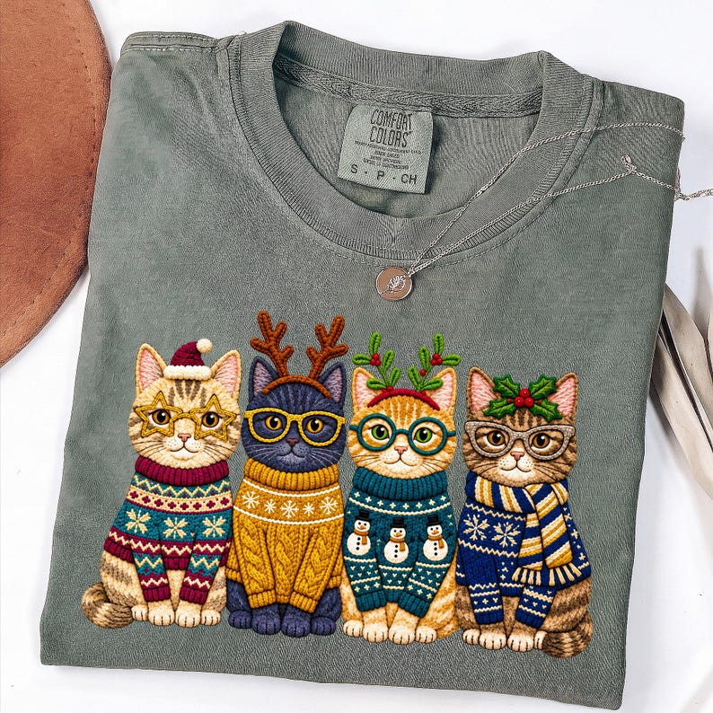 May include: A sage green t-shirt featuring a graphic of four cats wearing holiday sweaters, glasses, and festive headwear. The cats are dressed in patterned knitwear with reindeer antlers, holly, and Santa hats. The shirt has a Comfort Colors label.
