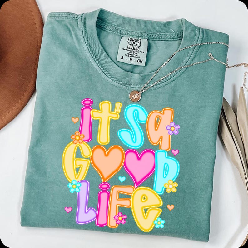 Pode incluir: Uma camiseta Comfort Colors verde-azulado com a frase "It's a Good Life" em letras coloridas. O design inclui cora&ccedil;&otilde;es e flores. Um colar de prata com um pingente est&aacute; sobre a camiseta.