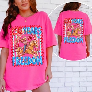 May include: Bright pink t-shirt with a graphic featuring a cowgirl riding a horse, lasso in hand, with the text "250 YEARS OF FREEDOM." The design is repeated on the back. The shirt has a relaxed fit.
