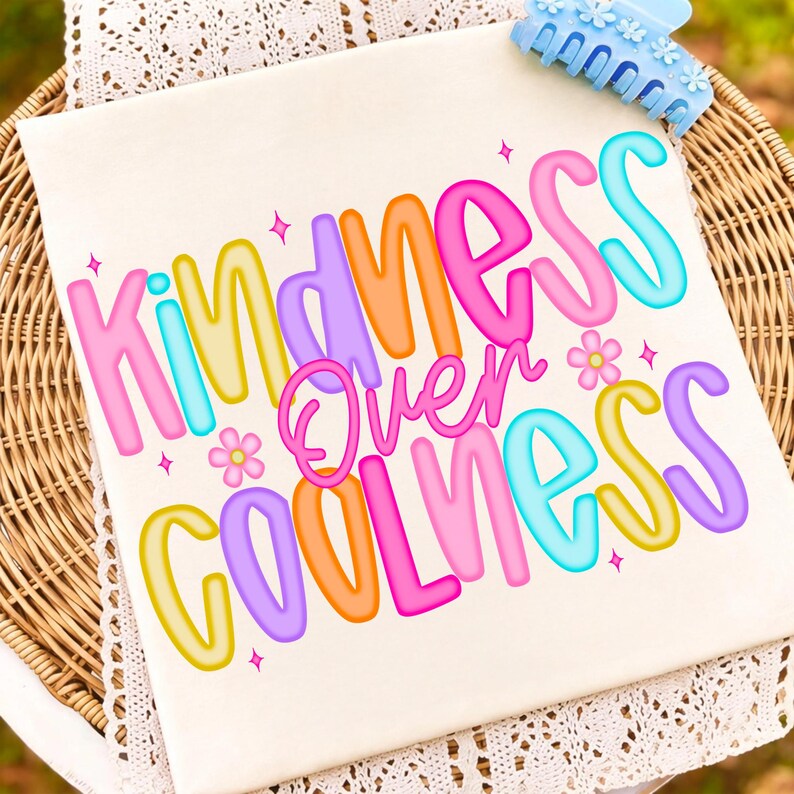 Może przedstawiać: Kremowa koszulka z napisem "KINDNESS Over COOLNESS" w kolorowych literach bąbelkowych. Projekt zawiera kwiaty i gwiazdki. Jasnoniebieska spinka do włos&oacute;w z kwiatowymi detalami znajduje się w prawym g&oacute;rnym rogu.