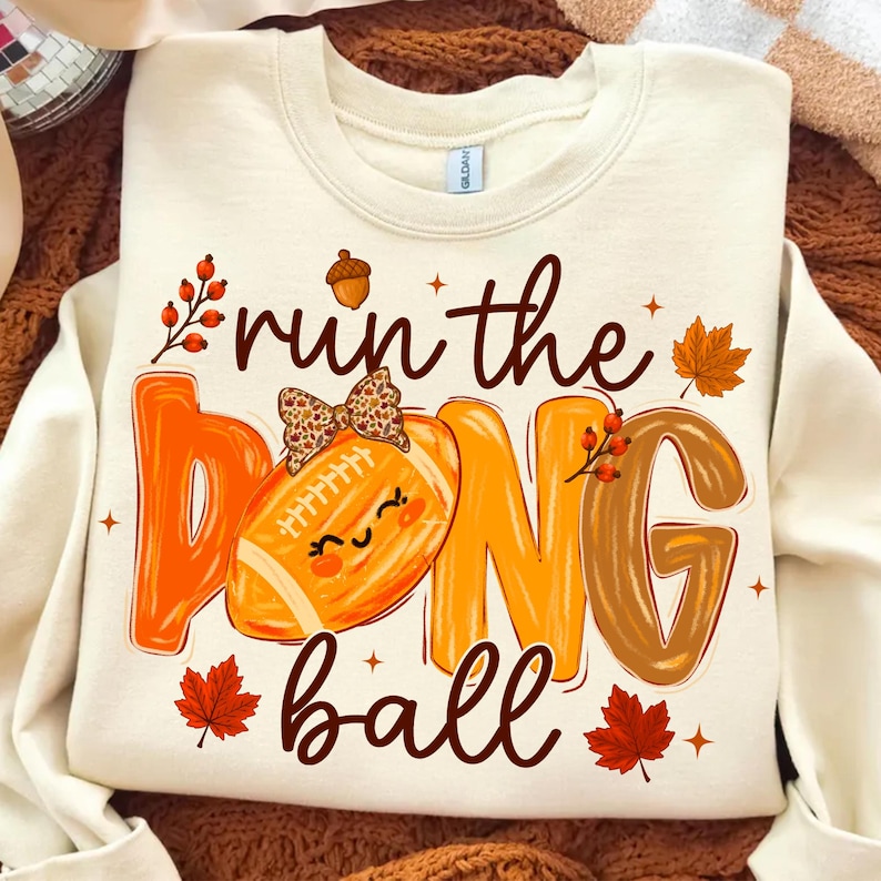 A close-up of a t-shirt featuring the 'Retro Football Mom Run The Dang Ball' PNG design with a coquette bow, showcasing vibrant autumn colors.