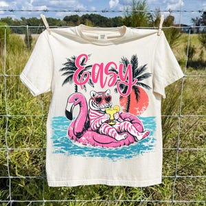 K&ouml;nnte beinhalten: Cremefarbenes T-Shirt mit einem Aufdruck eines Tigers mit Sonnenbrille, der auf einem rosa Flamingo-Schwimmer liegt und ein Getr&auml;nk h&auml;lt. Das Wort "Easy" steht in rosa Schrift dar&uuml;ber. Palmen und ein Sonnenuntergang sind im Hintergrund.