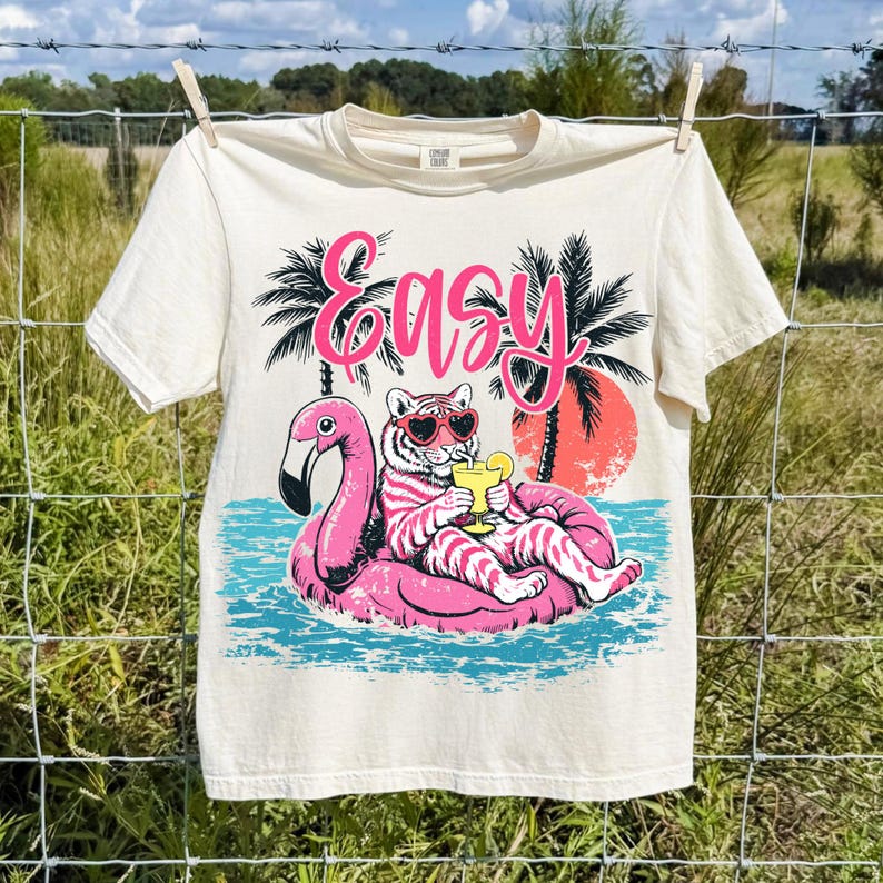 K&ouml;nnte beinhalten: Cremefarbenes T-Shirt mit einem Aufdruck eines Tigers mit Sonnenbrille, der auf einem rosa Flamingo-Schwimmer liegt und ein Getr&auml;nk h&auml;lt. Das Wort "Easy" steht in rosa Schrift dar&uuml;ber. Palmen und ein Sonnenuntergang sind im Hintergrund.