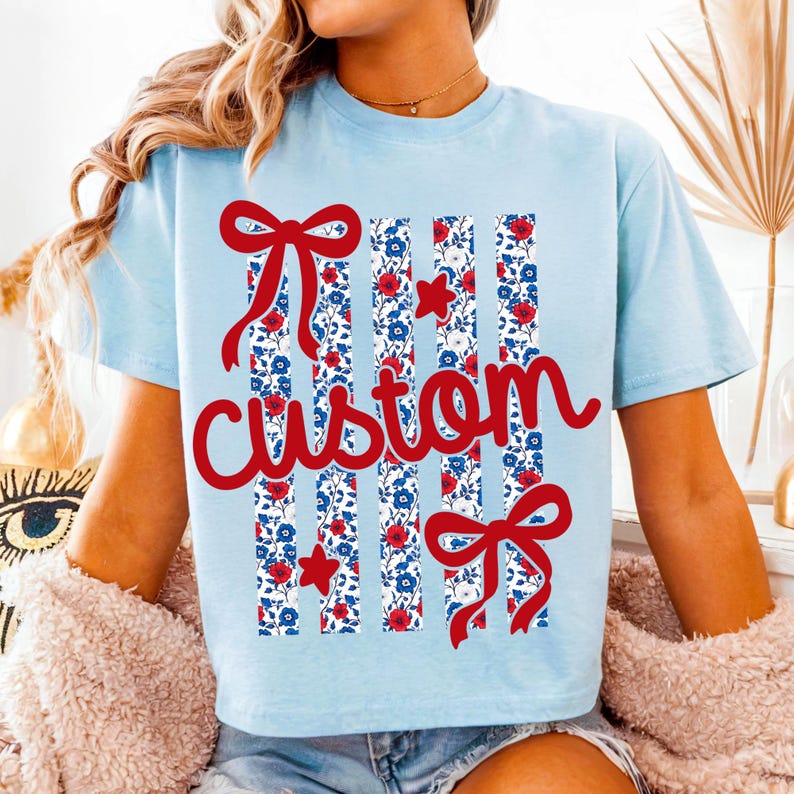 Puede incluir: Camiseta azul claro con un dise&ntilde;o "custom" rojo. El dise&ntilde;o incluye lazos rojos, estrellas rojas y rayas verticales con un estampado floral en rojo, blanco y azul. La camiseta tiene mangas cortas.