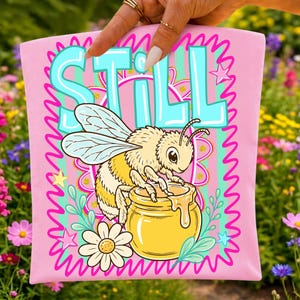 Op de afbeelding: Roze t-shirt met het woord "STILL" in turquoise letters. Een cartoonbij zit op een honingpot, met een madeliefje en sterren. Het ontwerp is omlijnd met een golvende roze rand.