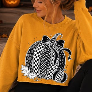 K&ouml;nnte beinhalten: Ein senfgelbes Langarm-Sweatshirt mit Rundhalsausschnitt und einem gro&szlig;en schwarz-wei&szlig;en K&uuml;rbis-Aufdruck, der mit einer Schleife verziert ist. Das K&uuml;rbis-Design zeigt Karomuster, Blattmotive, Eicheln und Sterne sowie zwei Eichenbl&auml;tter am unteren Rand.
