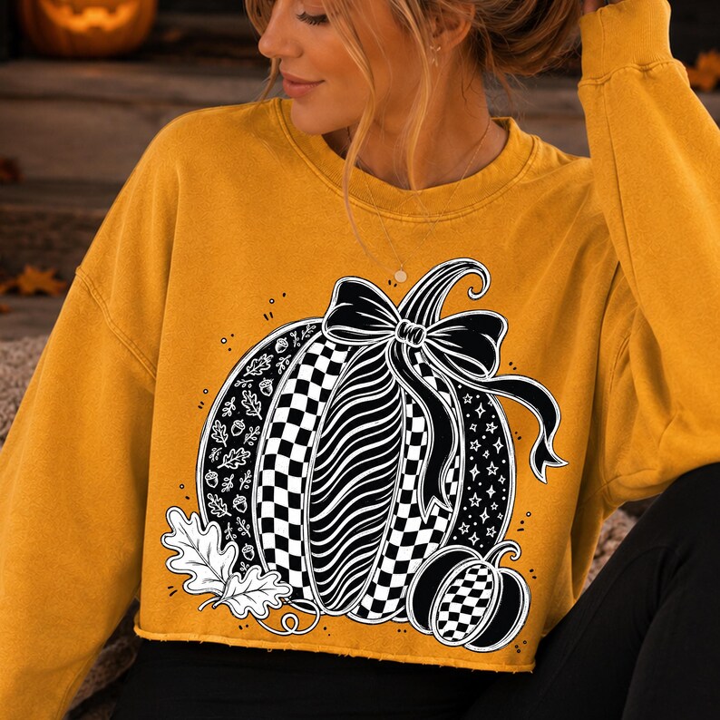 K&ouml;nnte beinhalten: Ein senfgelbes Langarm-Sweatshirt mit Rundhalsausschnitt und einem gro&szlig;en schwarz-wei&szlig;en K&uuml;rbis-Aufdruck, der mit einer Schleife verziert ist. Das K&uuml;rbis-Design zeigt Karomuster, Blattmotive, Eicheln und Sterne sowie zwei Eichenbl&auml;tter am unteren Rand.