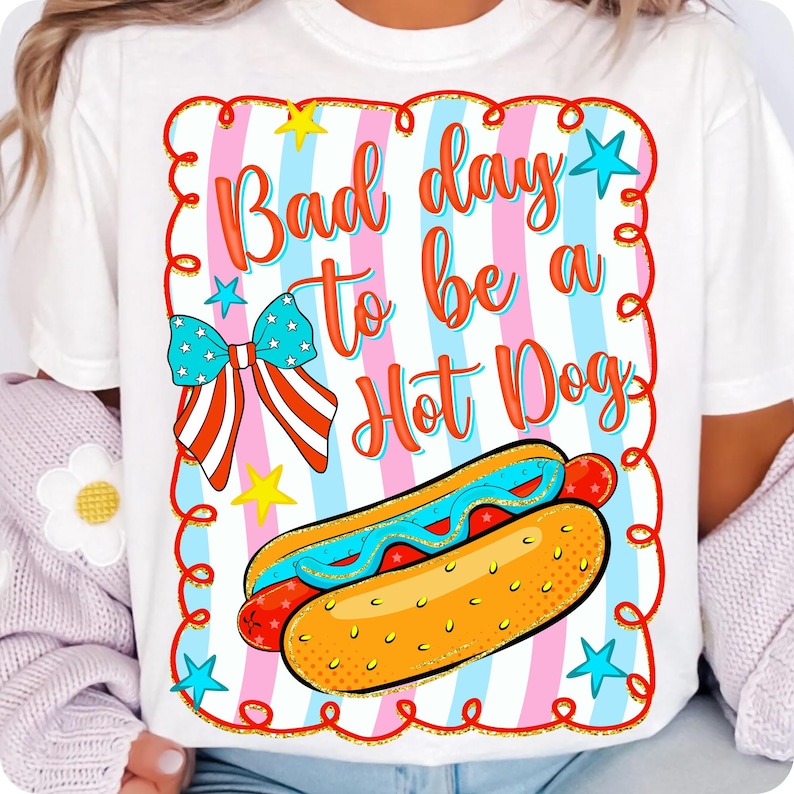 May include: White t-shirt with a colorful graphic that says "Bad day to be a Hot Dog." The design includes a hot dog, a bow, stars, and a decorative border. The shirt has a casual, relaxed fit.