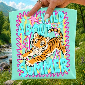 K&ouml;nnte beinhalten: Eine hellt&uuml;rkisfarbene Tragetasche mit einem Zeichentricktiger mit orangefarbenen und schwarzen Streifen. Die Tasche zeigt die Worte "WILD ABOUT SUMMER" in einer verspielten Schriftart, umgeben von Sternen und wellenf&ouml;rmigen Linien.