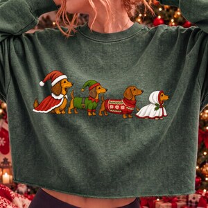 May include: A dark green long-sleeve sweatshirt featuring a graphic print of four dachshunds dressed in holiday attire. The dogs are wearing a Santa hat and cape, an elf costume, a red patterned jumper, and a white hooded cloak with holly.